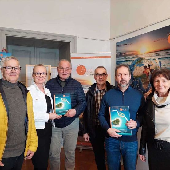 Eine Gruppe von Menschen steht lächelnd in einem Raum und hält Bücher. Der Mann in der Mitte trägt eine Brille und eine Jacke. Hinter ihnen ein Poster mit einem Globus und einem Sonnenuntergang.