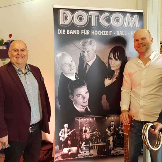 Zwei Männer stehen neben einem Poster der Band Dotcom, das ein Bandmitglied beim Gitarrespielen und andere bei Auftritten auf der Bühne zeigt.