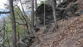 Ein steiler, felsiger Hang mit verstreuten Blättern und Bäumen, mit einem schmalen Pfad, der hinaufführt. Das Gelände ist uneben, und die Felsen sind zerklüftet.