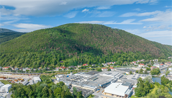 Luftaufnahme eines bewaldeten Berges mit einem großen Industriekomplex darunter. Der Berg ist grün mit einigen braunen Flecken, und der Komplex besteht aus mehreren Gebäuden mit flachen Dächern.