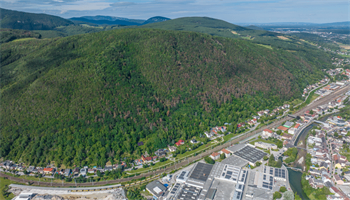 Ein Luftbild einer bewaldeten Anhöhe mit einigen Bäumen, die Anzeichen von Dürre zeigen, umgeben von einer kleinen Stadt mit Gebäuden und einem Fluss.