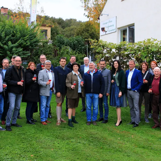 Eine Gruppe von Erwachsenen steht vor einem Gebäude mit dem Schriftzug 'Weinberg House'. Sie halten Weingläser in der Hand, wahrscheinlich bei einer gesellschaftlichen Veranstaltung. Bäume und Büsche umgeben das Gelände.