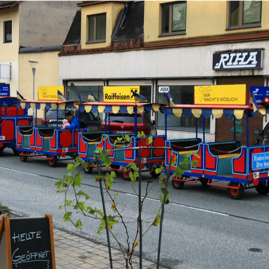 Vier bunte Spielzeugzüge sind am Straßenrand geparkt. Eine Person sitzt in einem der Züge. Die Straße hat eine Tafel mit deutschem Text. Hinter den Zügen befindet sich ein Gebäude mit einem Schild, auf dem 'RIFA' steht.