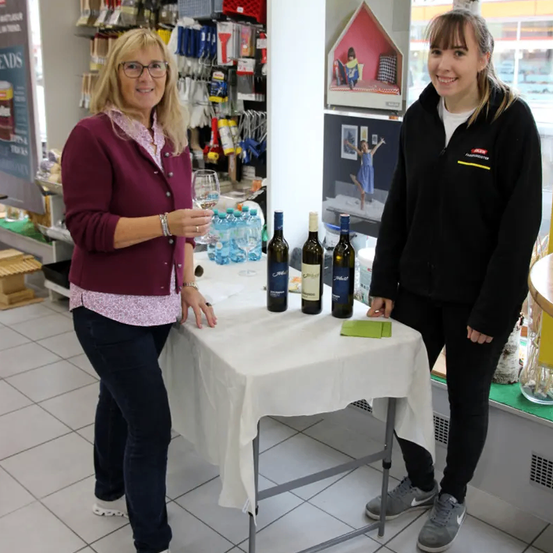 Zwei Frauen stehen neben einem Tisch mit Weinflaschen. Eine hält ein Weinglas, die andere lächelt. Sie befinden sich in einem Geschäft mit Regalen voller Artikel im Hintergrund.