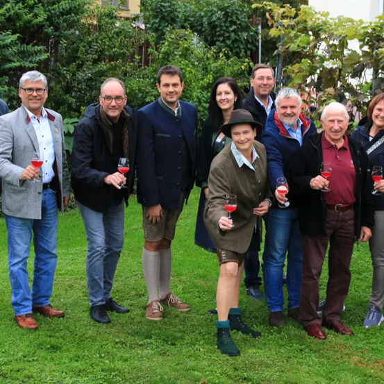 Eine Gruppe von Menschen steht in einem Garten, lächelt und hält Weingläser. Eine Frau in der Mitte trägt einen Hut und hält ein Weinglas mit rotem Wein.