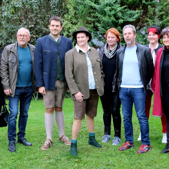 Eine Gruppe von Menschen, einige in traditioneller Kleidung, posiert für ein Foto auf einer Rasenfläche. Bäume und Büsche sind im Hintergrund zu sehen.