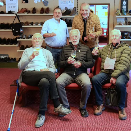 Fünf Männer sitzen auf einer roten Bank, drei halten Weingläser in der Hand, lächeln und posieren für ein Foto. Dahinter sind Regale mit Schuhen gefüllt.