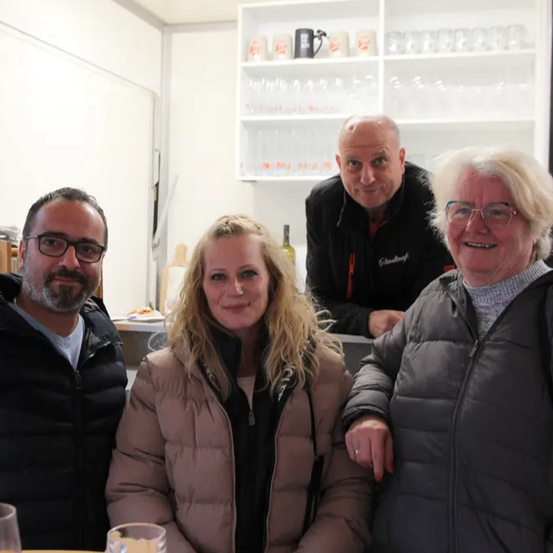 Vier Menschen, zwei Männer und zwei Frauen, lächeln für ein Foto. Sie tragen Jacken und Brillen. Die Frau in der Mitte hat blonde Haare. Hinter ihnen steht ein Mann in der Nähe eines Regals.