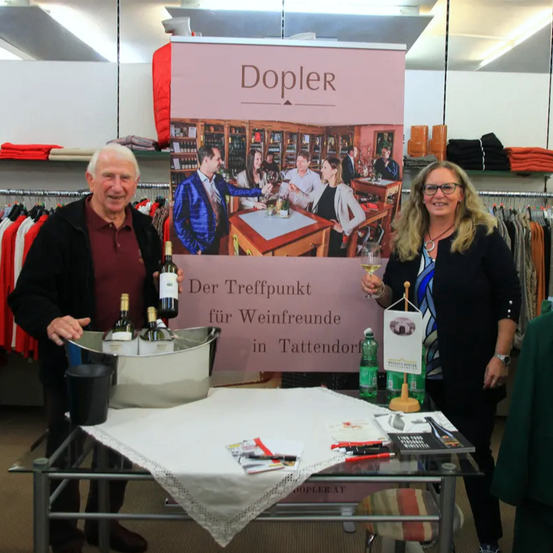 Zwei Personen stehen vor einem Tisch mit Weinflaschen in einem Eiskübel. Ein Schild hinter ihnen lautet 'Der Treffpunkt für Weinfreunde in Tattendorf.' Sie lächeln und halten Weingläser in den Händen.