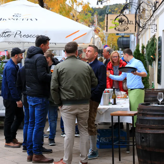 Eine Gruppe von Männern und Frauen steht draußen in einer festlichen Umgebung, vielleicht auf einem Weinfest. Ein Mann hält einen Tablett mit Weingläsern. Ein Zeltschirm ist in der Nähe, mit einem Schild, das Monetto Prosecco anzeigt.