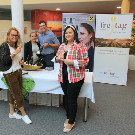 Eine Gruppe von vier Personen steht hinter einem Tisch mit Weinflaschen. Zwei Frauen halten Weingläser und ein Mann hält eine Flasche. Die Frau in der Mitte trägt ein kariertes Hemd. Dahinter befindet sich ein Banner mit dem Text 'freytag'.