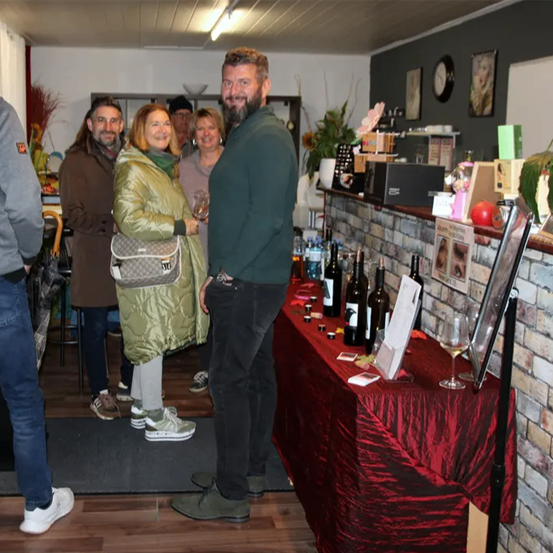 Mehrere Personen sind in einem Raum versammelt. Ein Mann in der Mitte hält ein Weinglas. Hinter ihm steht ein Tisch, der mit einem roten Tuch bedeckt ist und verschiedene Flaschen zeigt. Eine Frau auf der linken Seite trägt eine Tasche. Der Raum hat eine Ziegelwand und ein Regal mit verschiedenen Gegenständen.