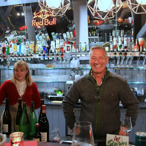 Ein lächelnder Mann und eine Frau stehen hinter einer Bar mit verschiedenen Flaschen und Gläsern. Dahinter leuchtet ein Red Bull-Neon-Schild und Regale voller Alkoholflaschen.