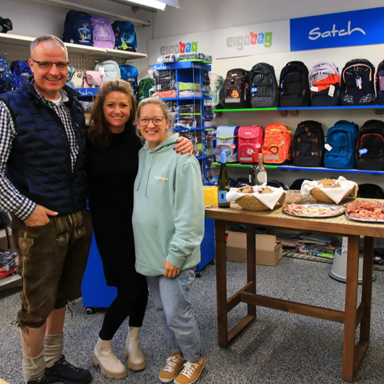 Drei Personen stehen in einem Geschäft mit Rucksackregalen im Hintergrund. Ein Tisch hält ein Fleischteller, Weinflaschen und Körbe. Der Mann trägt eine Brille und eine Weste, die Frau links Stiefel und die Frau rechts eine Kapuzenjacke.