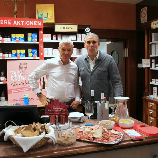 Zwei Männer stehen hinter einer Theke in einem Geschäft. Der Mann auf der linken Seite trägt ein weißes Hemd und hat ein Schild hinter sich, auf dem 'Der Rose Bauer' steht. Verschiedene Lebensmittel und Getränke stehen auf der Theke, zusammen mit einem Monitor und einem Regal, das mit Produkten gefüllt ist.