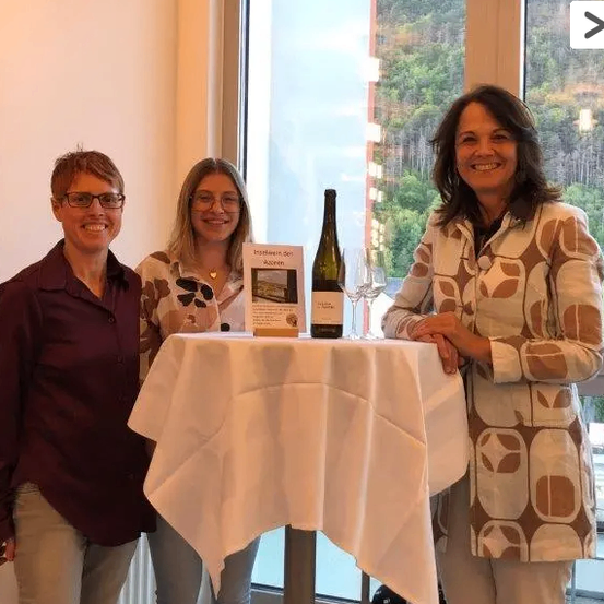 Drei Frauen stehen lächelnd neben einem Tisch mit einer Flasche Wein und Weingläsern, vor einem malerischen Ausblick durch ein Fenster.
