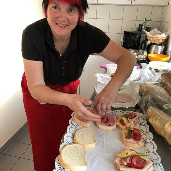 Eine Frau mit roten Haaren, die ein schwarzes Shirt und einen roten Rock trägt, bereitet Sandwiches auf einem Teller mit einer Spitzendeckchen in einer Küche zu.