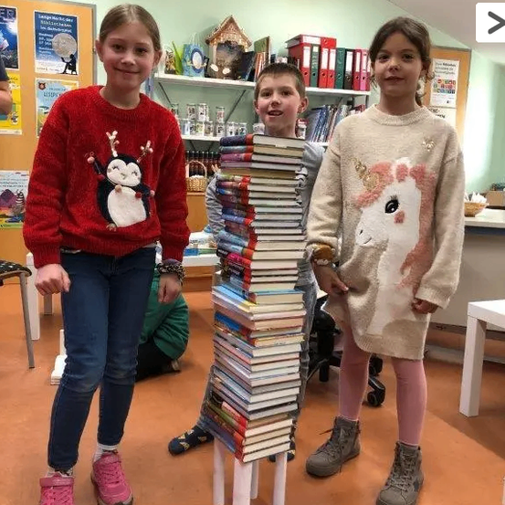 Drei Kinder stehen lächelnd in einer Bibliothek. Ein Mädchen trägt ein Rentier-Sweatshirt, ein Junge ein Pinguin-Sweatshirt und ein anderes Mädchen ein Einhorn-Sweatshirt. Sie posieren neben einem hohen Stapel Bücher.