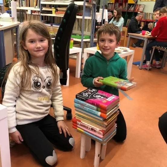 Zwei Kinder posieren für ein Foto in einer Bibliothek, eines hält einen Stapel Bücher. Das Mädchen trägt einen Pullover mit Eulen-Design.