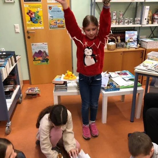 Ein kleines Mädchen steht mit erhobenen Armen in einer Bibliothek, während Kinder um sie herum sitzen und lesen. Bücher, Spielzeug und Poster schmücken den Raum.