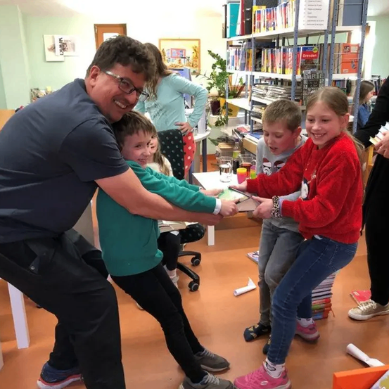 Ein Mann mit Brille hält die Hände zweier Kinder, eines in Grün und eines in Rot, während sie alle lächeln. Hinter ihnen sehen eine Frau und ein weiteres Kind zu. Ein Tisch mit Büchern und Gegenständen ist im Vordergrund, mit Regalen und einer Pflanze im Hintergrund.