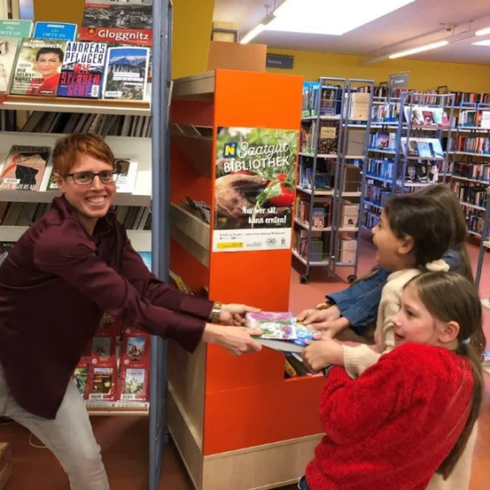 Eine Frau mit Brille gibt zwei jungen Mädchen Bücher in einer Bibliothek. Hinter ihnen Bücherregale und ein Schild mit der Aufschrift 'Saatgut Bibliothek'.