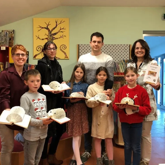 Eine Gruppe von Kindern und Erwachsenen posiert für ein Foto in einer Bibliothek, wobei sie Bücher mit einzigartigen Designs in den Händen halten. Dahinter zeigt eine Wand ein großes, gelbes Baumgemälde.