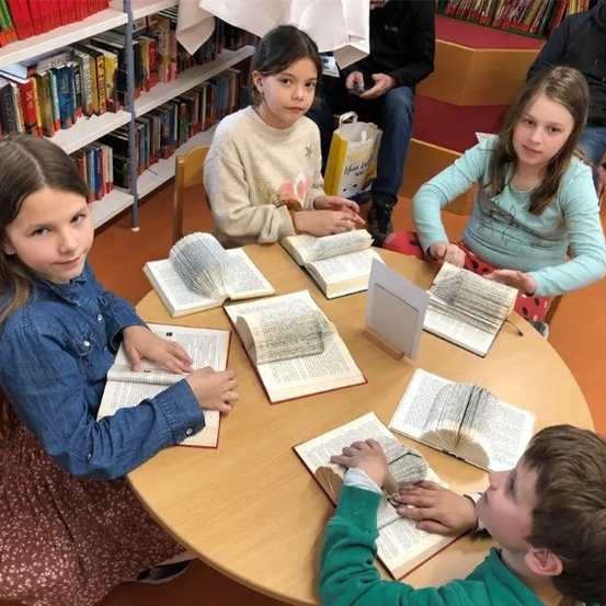 Fünf Kinder sitzen in einer Bibliothek um einen Tisch herum, jedes hält ein offenes Buch. Der Tisch ist mit mehreren Büchern bedeckt. Dahinter befinden sich Regale voller Bücher und eine Person, die auf einer Bank sitzt.