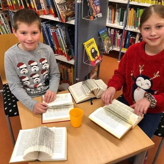 Zwei Kinder sitzen an einem Tisch in einer Bibliothek, jedes mit einem offenen Buch. Ein Junge hat ein Buch in seinem Schoß, während ein Mädchen ein offenes Buch hält. Sie sind von Büchern und Regalen umgeben.