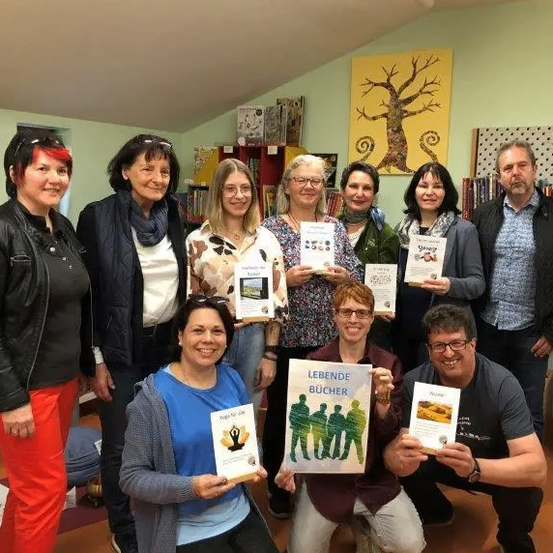 Eine Gruppe Erwachsener posiert für ein Foto in einem Raum mit Büchern und einem Baumbild an der Wand. Sie halten alle Bücher in den Händen, und einige tragen Brillen.