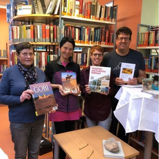 Vier Personen posieren für ein Foto in einer Bibliothek, jeder hält ein Buch in der Hand, mit Bücherregalen voller Bücher im Hintergrund.