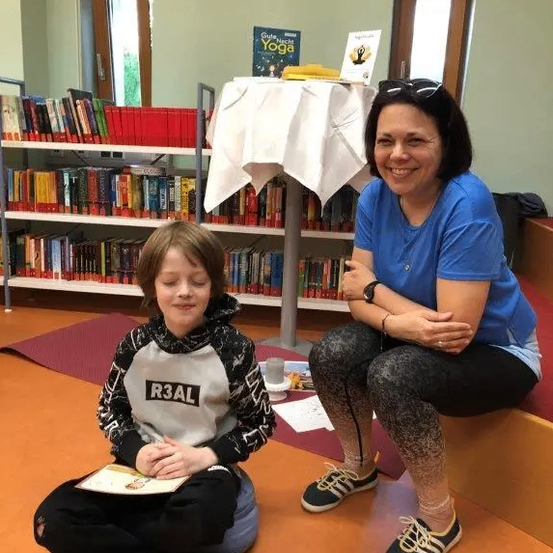 Eine erwachsene Frau und ein junger Junge sitzen auf einer Bank in einer Bibliothek. Der Junge hält ein Buch in der Hand und lächelt. Die Frau trägt eine Sonnenbrille auf dem Kopf und eine Halskette.
