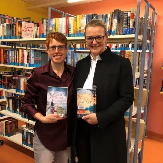 Zwei Frauen stehen in einer Bibliothek, lächeln und halten Bücher in den Händen. Ein Buch heißt 'Sturm Madchen' und das andere 'Tee Palast'. Regale voller Bücher stehen hinter ihnen.