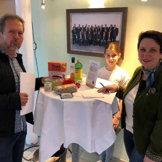 Drei Personen stehen um einen Tisch mit Essensartikeln und einem Schild. Ein Mann hält ein Papier, eine Frau eine Feder und ein Mädchen lächelt.