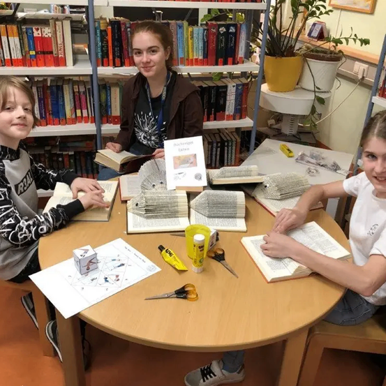 Drei Kinder sitzen um einen Tisch herum, jedes mit einem Buch und Bastelwerkzeugen. Der Tisch ist mit Büchern und Bastelutensilien beladen. Eine Frau steht hinter ihnen und hält ein Buch mit dem Titel 'Bucherbommel'. Der Raum hat Bücherregale und Topfpflanzen.
