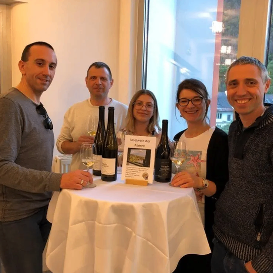 Fünf Personen stehen um einen Tisch mit Weinflaschen und Gläsern und lächeln für ein Foto. Hinter ihnen ist ein Fenster mit Blick auf Häuser und Bäume.