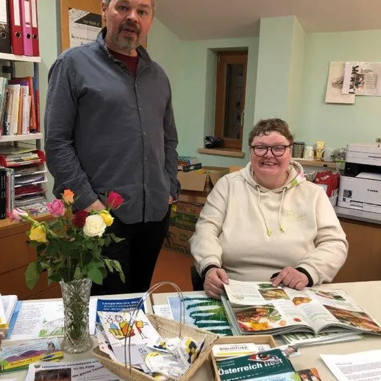 Ein Mann und eine Frau sitzen an einem Tisch mit Büchern, einer Blumenvase und anderen Gegenständen. Dahinter steht ein Bücherregal mit Büchern und Ordnern. Rechts steht ein Drucker.