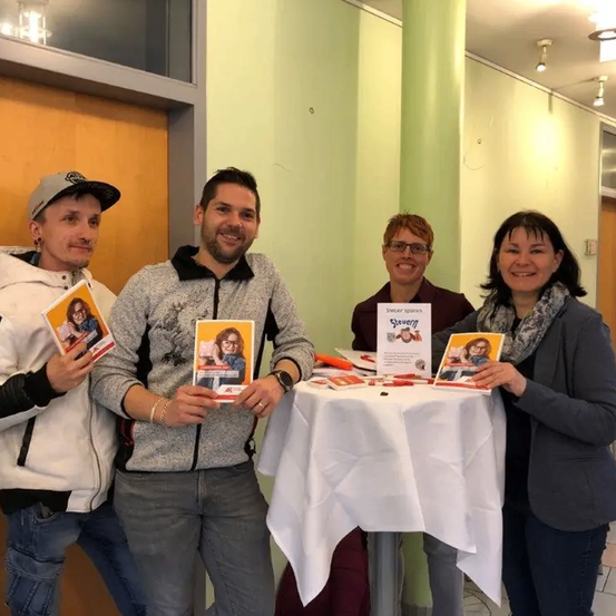 Fünf Personen stehen hinter einem Tisch mit Büchern in der Hand. Sie lächeln und scheinen für ein Foto zu posieren. Jede Person hält ein Buch mit einem Bild einer Frau. Der Tisch ist mit einem weißen Tuch bedeckt. Dahinter befindet sich eine Wand mit einer grünen Säule.