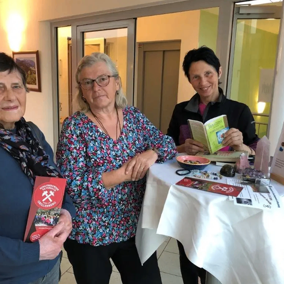 Drei Frauen stehen hinter einem Tisch mit Büchern und Steinen. Die Frau auf der linken Seite hält ein rotes Buch. Die Frau in der Mitte trägt eine Brille und eine Bluse mit Blumenmuster. Die Frau auf der rechten Seite hält ein Buch und eine Schüssel.