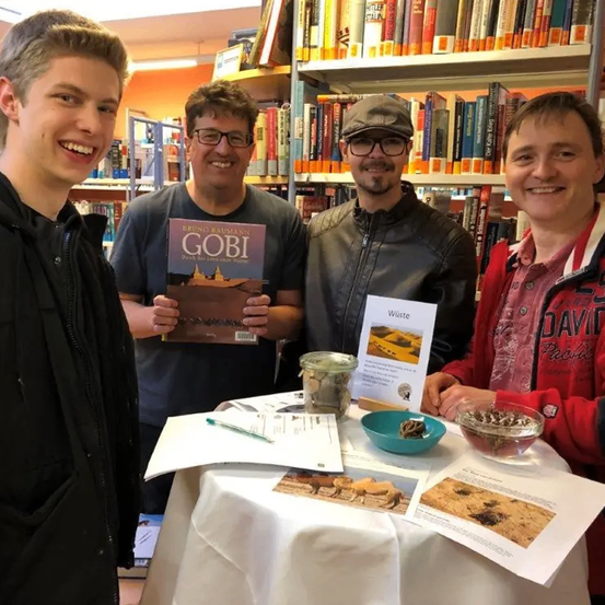 Vier Männer posieren in einer Bibliothek mit Büchern und einem Tisch mit Gegenständen, lächeln und schauen in die Kamera.