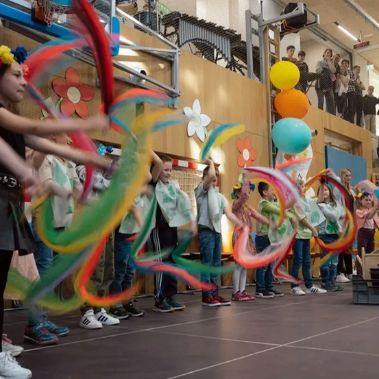 Eine Gruppe von Kindern führt eine Tanzroutine mit bunten Bändern auf einer Bühne auf, Zuschauer beobachten vom Balkon.