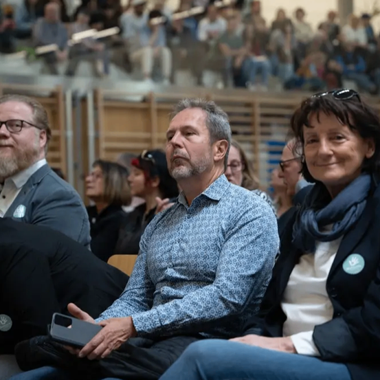 Eine Gruppe von Menschen sitzt in Stühlen nach vorne gerichtet, einige mit Brille. Ein Mann in einem blauen Hemd hält ein Telefon. Eine Frau in einer blauen Jacke lächelt.