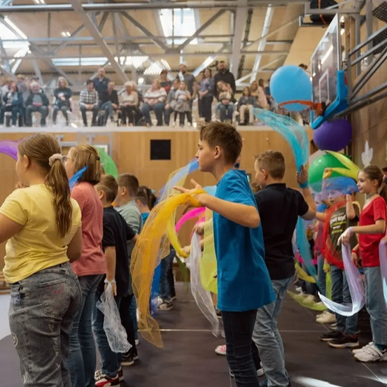 Kinder führen mit bunten Bändern in einer großen Halle auf, beobachtet von einem Publikum auf der Empore.
