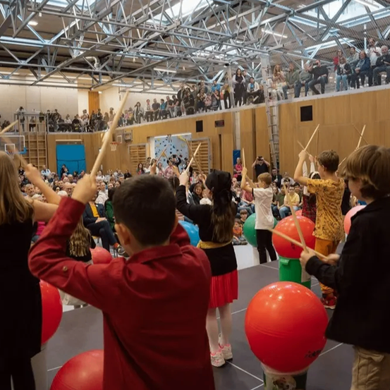 Eine Gruppe von Kindern spielt Trommeln auf einer Bühne vor einem Publikum. Die Kinder halten Trommelstöcke und die Bühne ist von roten Bällen umgeben.