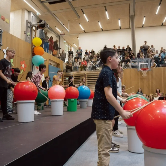 Eine Gruppe von Kindern spielt Trommeln auf einer Bühne vor einem Publikum. Die Trommeln sind groß und farbenfroh, einige auf Eimern. Die Kinder lächeln und scheinen sich zu amüsieren.