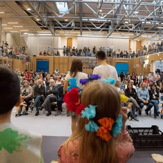 Eine Gruppe von Menschen schaut sich eine Aufführung in einer großen Halle an. Einige sitzen auf Tribünen, andere stehen. Ein Mädchen in einem rosa Kleid hat bunte Blumen in den Haaren.
