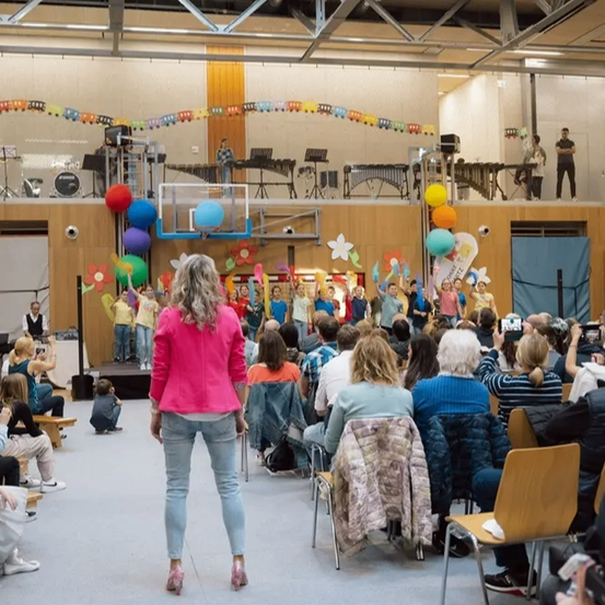 Eine Frau in einer rosa Jacke steht vor einem sitzenden Publikum in einem dekorierten Saal. Musikinstrumente stehen auf der Bühne, und Ballons hängen. Leute machen Fotos.