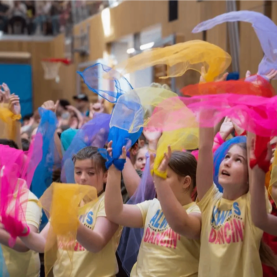 Eine Gruppe von Menschen in gelben T-Shirts schwenkt bunte Bänder in einer Turnhalle, einige tragen Brillen und Armbänder. Sie nehmen wahrscheinlich an einer Tanzveranstaltung teil.