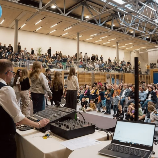 Ein Mann bedient eine Mischkonsole in einer Halle mit vielen Menschen. Das Publikum sitzt auf Bänken, einige stehen. Ein großer blauer Ballon hängt von der Decke.