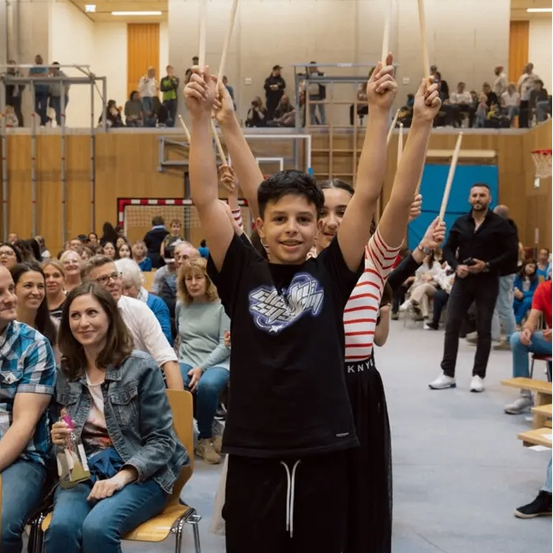 Ein Junge hebt seine Hände in einer überdachten Sporthalle, möglicherweise einem Basketballplatz, während um ihn herum Menschen sitzen. Einige lächeln, andere schauen zu. Ein Mann in einem schwarzen Anzug steht vor den sitzenden Menschen. Der Bereich hat Holzbarrieren und einen Basketballkorb.
